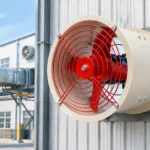 Industrial exhaust fan with red guard grille mounted on the corrugated steel wall of a factory building